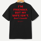 A black short-sleeve t-shirt hanging on a wooden hanger, showing the back with bold red text that reads 'I'M FRIENDLY, BUT MY WIFE ISN'T. Don't touch!' and a small red YLF logo below.