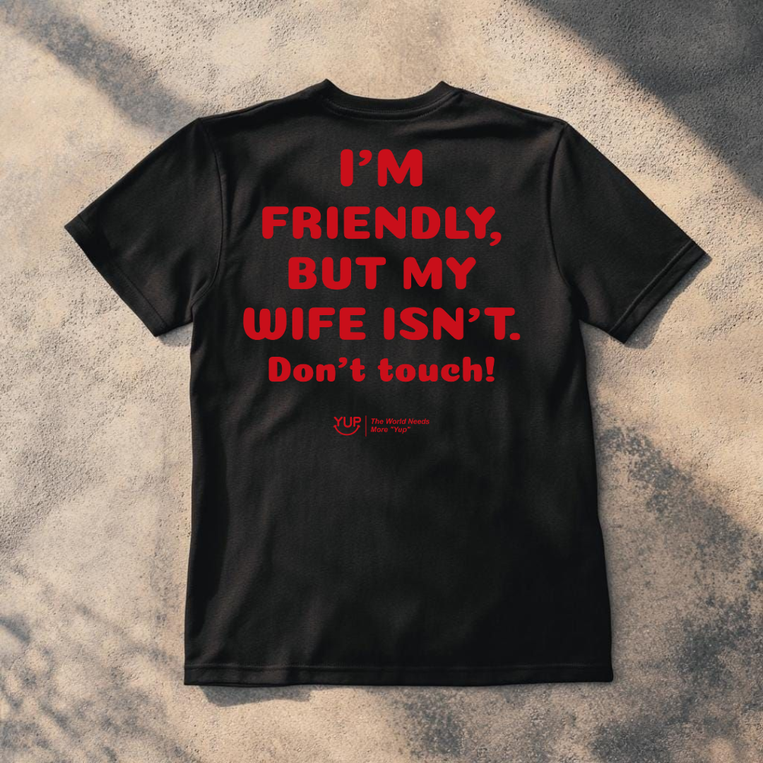 A black short-sleeve t-shirt laid flat on a textured concrete surface with dappled shadows. The back of the shirt features bold red text that reads 'I'M FRIENDLY, BUT MY WIFE ISN'T.
