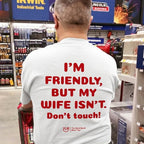 A man viewed from behind in a hardware store aisle, wearing a white t-shirt with bold red text that reads 'I'M FRIENDLY, BUT MY WIFE ISN'T. Don't touch!' on the back.