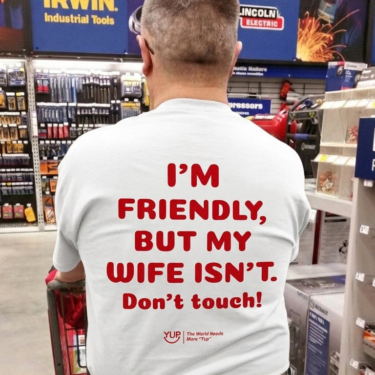 A man viewed from behind in a hardware store aisle, wearing a white t-shirt with bold red text that reads 'I'M FRIENDLY, BUT MY WIFE ISN'T. Don't touch!' on the back.