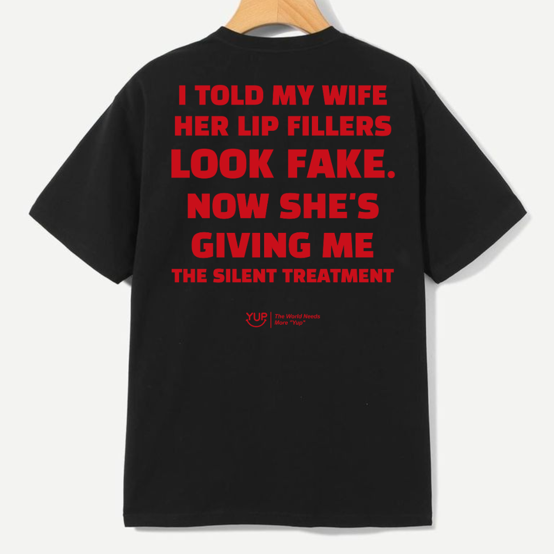 A black short-sleeve t-shirt hanging on a wooden hanger against a light gray background, featuring a humorous red text graphic on the back.
