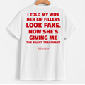 White short-sleeve T-shirt hanging on a wooden hanger against a light gray background, showing a large red text graphic on the back that reads, 'I TOLD MY WIFE HER LIP FILLERS LOOK FAKE. NOW SHE'S GIVING ME THE SILENT TREATMENT' with a small red YUP logo below.
