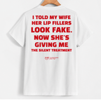 White short-sleeve T-shirt hanging on a wooden hanger against a light gray background, showing a large red text graphic on the back that reads, 'I TOLD MY WIFE HER LIP FILLERS LOOK FAKE. NOW SHE'S GIVING ME THE SILENT TREATMENT' with a small red YUP logo below.