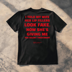 A black short-sleeve t-shirt laid flat on a textured concrete surface with dappled sunlight and shadows. The back of the shirt features a bold red text graphic that reads: 'I TOLD MY WIFE HER LIP FILLERS LOOK FAKE.