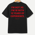 A black short-sleeve t-shirt hanging on a wooden hanger, showing the back with bold red text that reads 'I'M NOT 30. I'M 18 WITH 12 YEARS OF EXPERIENCE.