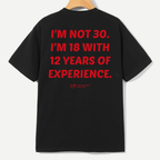 A black short-sleeve t-shirt hanging on a wooden hanger, showing the back with bold red text that reads 'I'M NOT 30. I'M 18 WITH 12 YEARS OF EXPERIENCE.