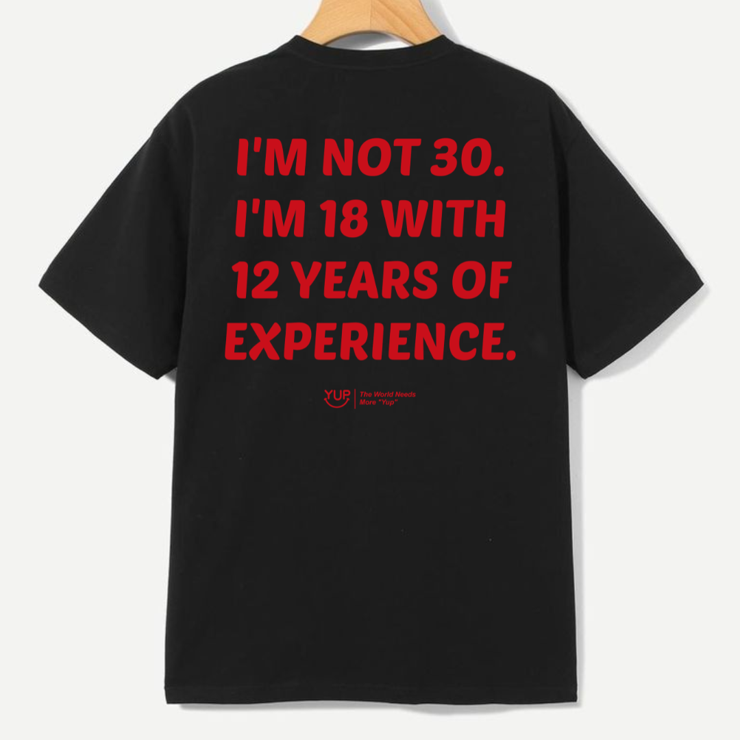A black short-sleeve t-shirt hanging on a wooden hanger, showing the back with bold red text that reads 'I'M NOT 30. I'M 18 WITH 12 YEARS OF EXPERIENCE.