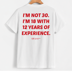 White short-sleeve T-shirt hanging on a wooden hanger against a light gray background, showing the back with bold red text that reads, 'I'M NOT 30. I'M 18 WITH 12 YEARS OF EXPERIENCE.