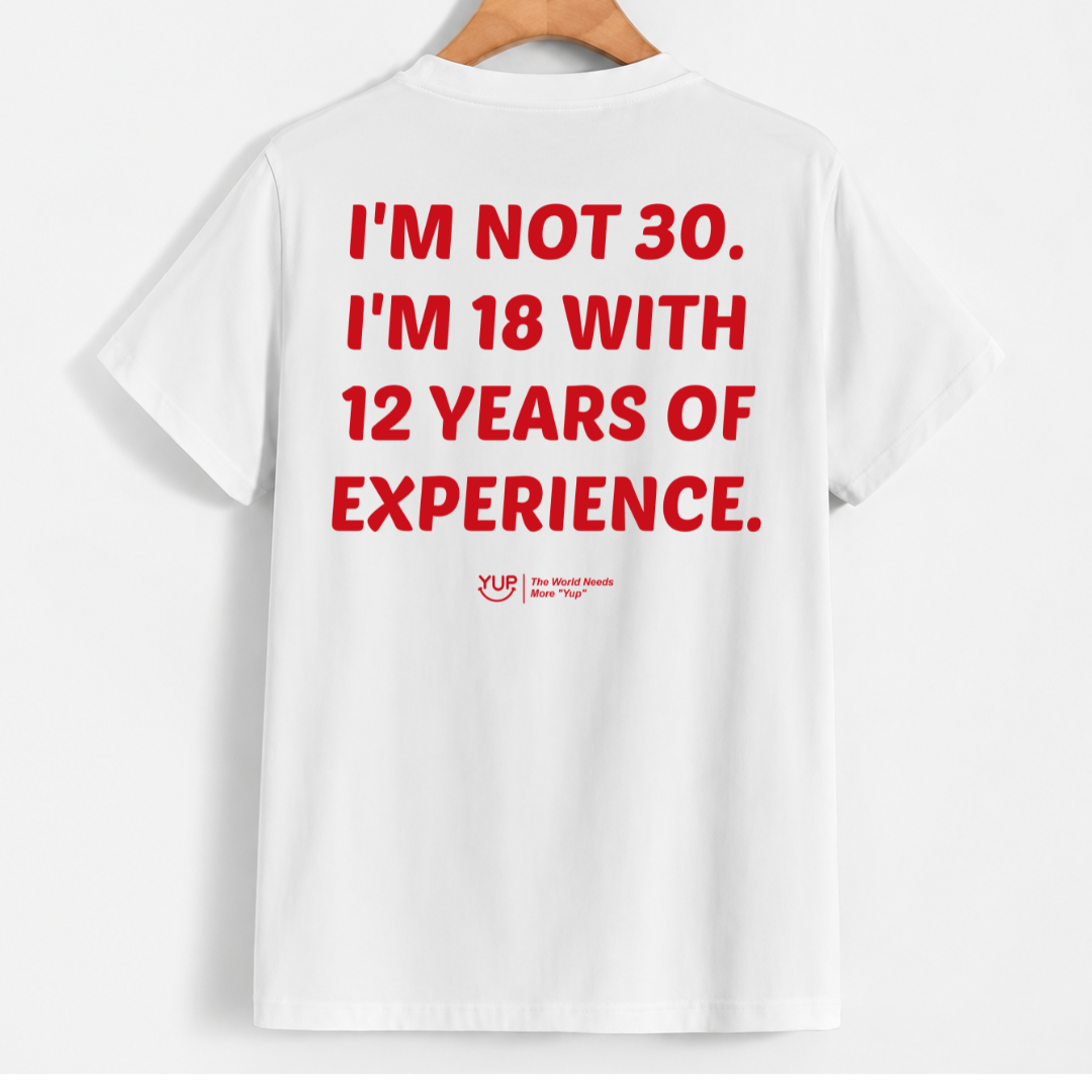 White short-sleeve T-shirt hanging on a wooden hanger against a light gray background, showing the back with bold red text that reads, 'I'M NOT 30. I'M 18 WITH 12 YEARS OF EXPERIENCE.