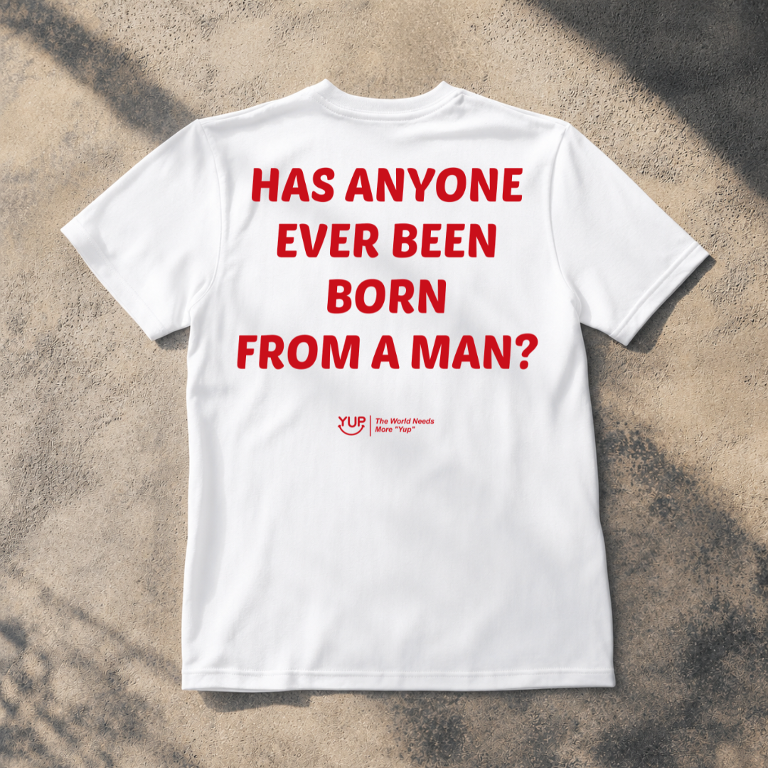 A white short-sleeve T-shirt laid flat on a textured concrete surface with shadows. The shirt has large red text reading 'HAS ANYONE EVER BEEN BORN FROM A MAN?' and a small red YUP logo and slogan near the lower center.