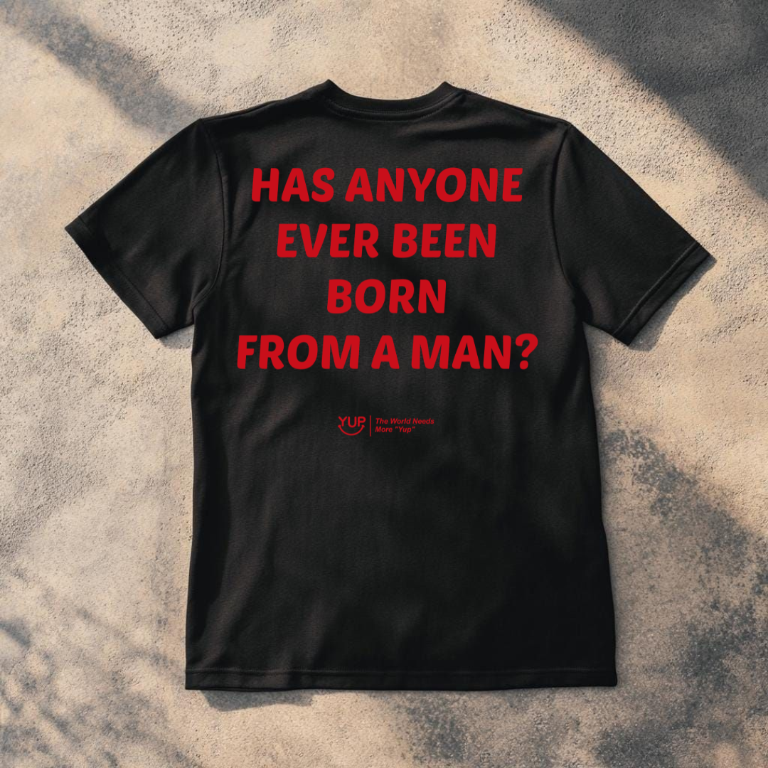 A black short-sleeve t-shirt laid flat on a textured concrete surface with dappled shadows. The back of the shirt features bold red text that reads 'HAS ANYONE EVER BEEN BORN FROM A MAN?' with a small red 'YUP' logo underneath.