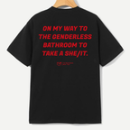 A black short-sleeve t-shirt hanging on a wooden hanger, showing the back with bold red text that reads 'ON MY WAY TO THE GENDERLESS BATHROOM TO TAKE A SHE/IT.' and a small red YUP logo underneath.