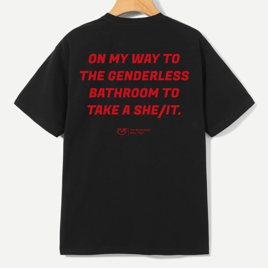 A black short-sleeve t-shirt hanging on a wooden hanger, showing the back with bold red text that reads 'ON MY WAY TO THE GENDERLESS BATHROOM TO TAKE A SHE/IT.' and a small red YUP logo underneath.