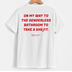 White short-sleeve T-shirt hanging on a wooden hanger against a light gray background, with bold red text on the back reading 'ON MY WAY TO THE GENDERLESS BATHROOM TO TAKE A SHE/IT.' and a small red YUP logo below.