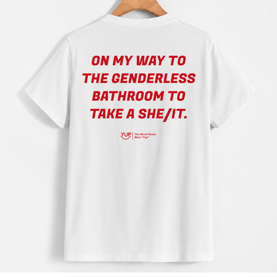 White short-sleeve T-shirt hanging on a wooden hanger against a light gray background, with bold red text on the back reading 'ON MY WAY TO THE GENDERLESS BATHROOM TO TAKE A SHE/IT.' and a small red YUP logo below.