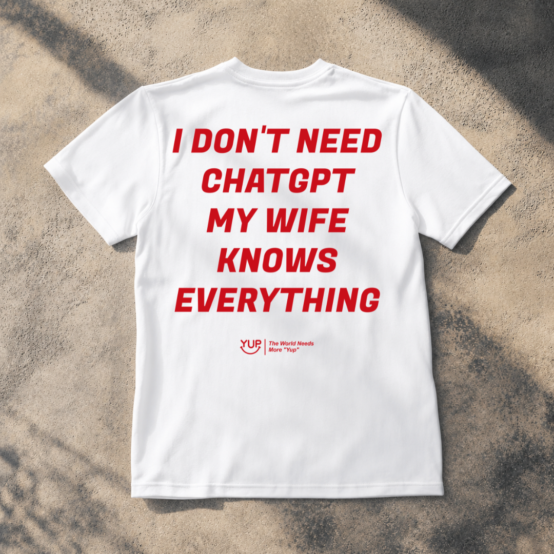 A white short-sleeve t-shirt laid flat on a textured concrete surface with dappled shadows. The shirt features bold red text reading 'I DON'T NEED CHATGPT MY WIFE KNOWS EVERYTHING' and a small red 'Yup The World Needs Me Be 'Yup!' logo underneath.