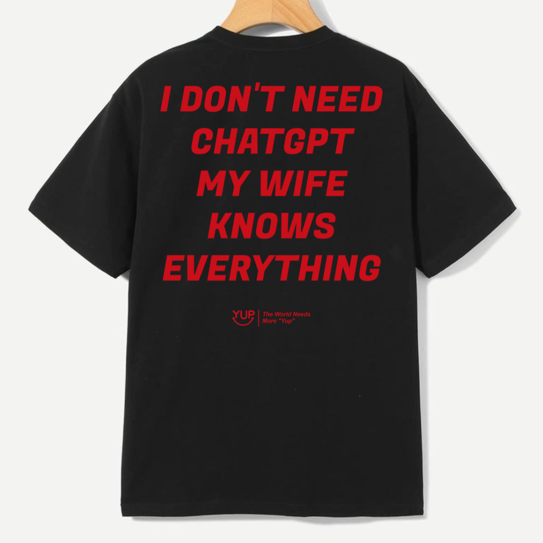 A black short-sleeve t-shirt hanging on a wooden hanger against a light gray background, featuring a bold red text graphic on the back that reads 'I DON'T NEED CHATGPT MY WIFE KNOWS EVERYTHING' with a small red 'Yupoong' logo underneath.