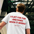 A man viewed from behind wearing a white t-shirt with bold red text that reads 'I FLIRT FOR FUN AND PANIC WHEN IT WORKS' and a small 'Yup! The World's Best' logo, paired with blue jeans in an indoor setting.