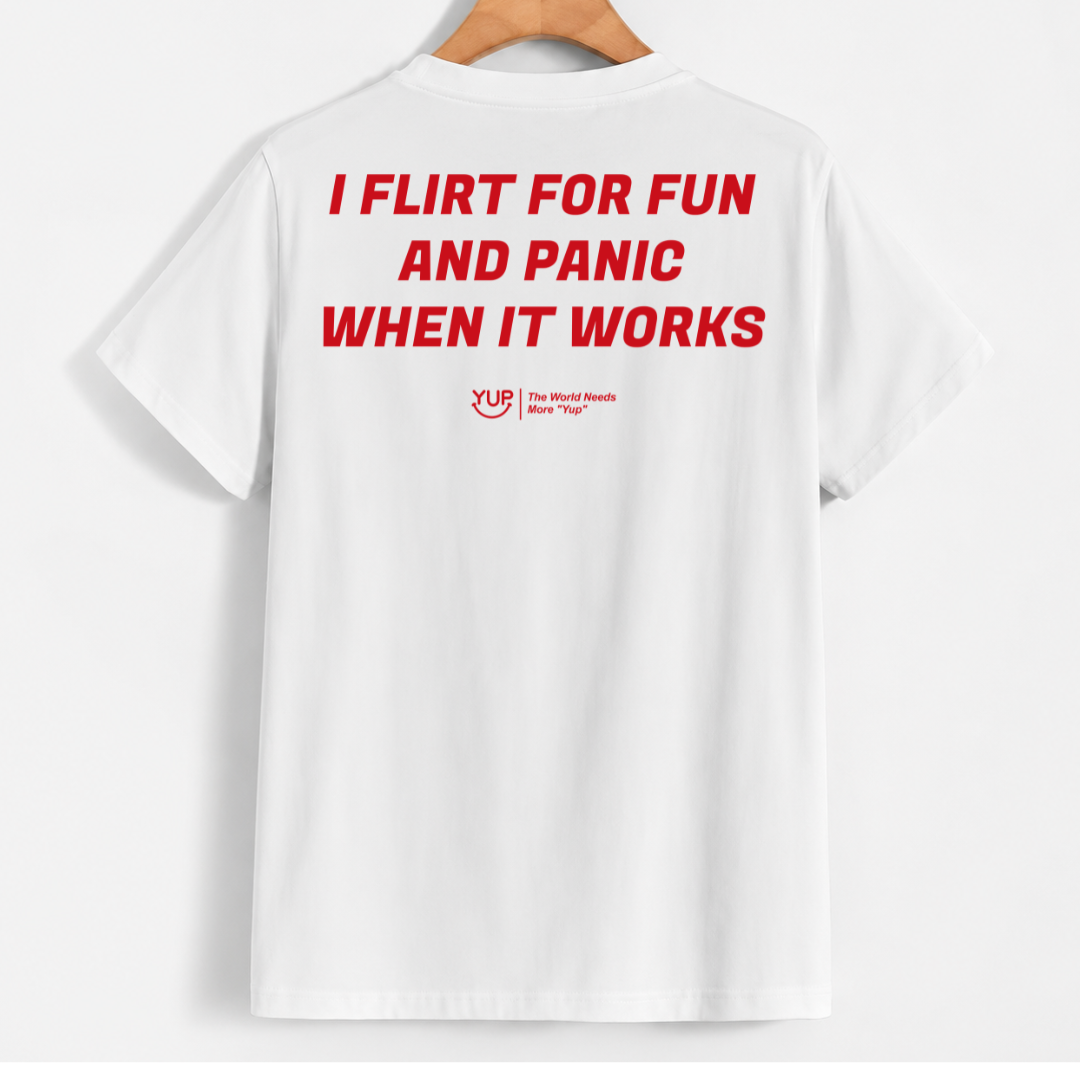 White short-sleeve t-shirt hanging on a wooden hanger, showing the back with bold red text that reads 'I FLIRT FOR FUN AND PANIC WHEN IT WORKS' and a small red 'YUS' logo underneath.
