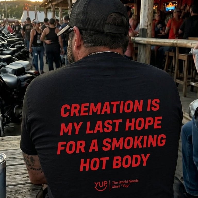 A man seen from behind wearing a black t-shirt with bold red text that reads 'CREMATION IS MY LAST HOPE FOR A SMOKING HOT BODY' on a wooden deck at an outdoor bar.