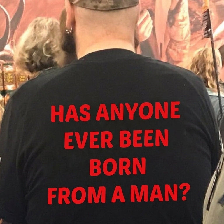 A man viewed from behind wearing a black t-shirt with bold red text that reads 'HAS ANYONE EVER BEEN BORN FROM A MAN?' and a small red logo near the bottom. He is wearing a camouflage cap in what appears to be a store or event setting.