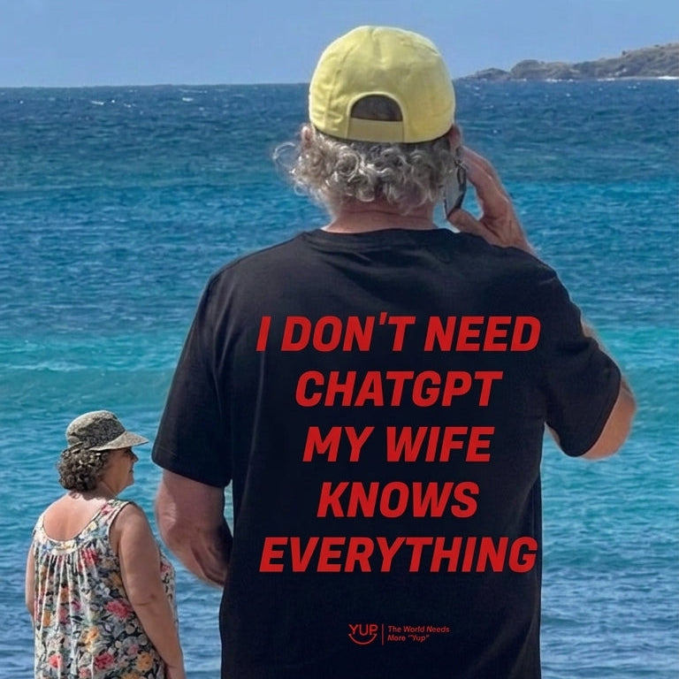 A man standing on a beach with his back to the camera, wearing a black t-shirt with bold red text that reads 'I DON'T NEED CHATGPT MY WIFE KNOWS EVERYTHING' and a yellow cap. A woman in a floral dress stands nearby looking at the ocean.