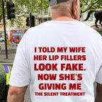 A man viewed from behind wearing a white t-shirt with bold red text printed on the back, standing on a city street. The text reads: 'I TOLD MY WIFE HER LIP FILLERS LOOK FAKE.