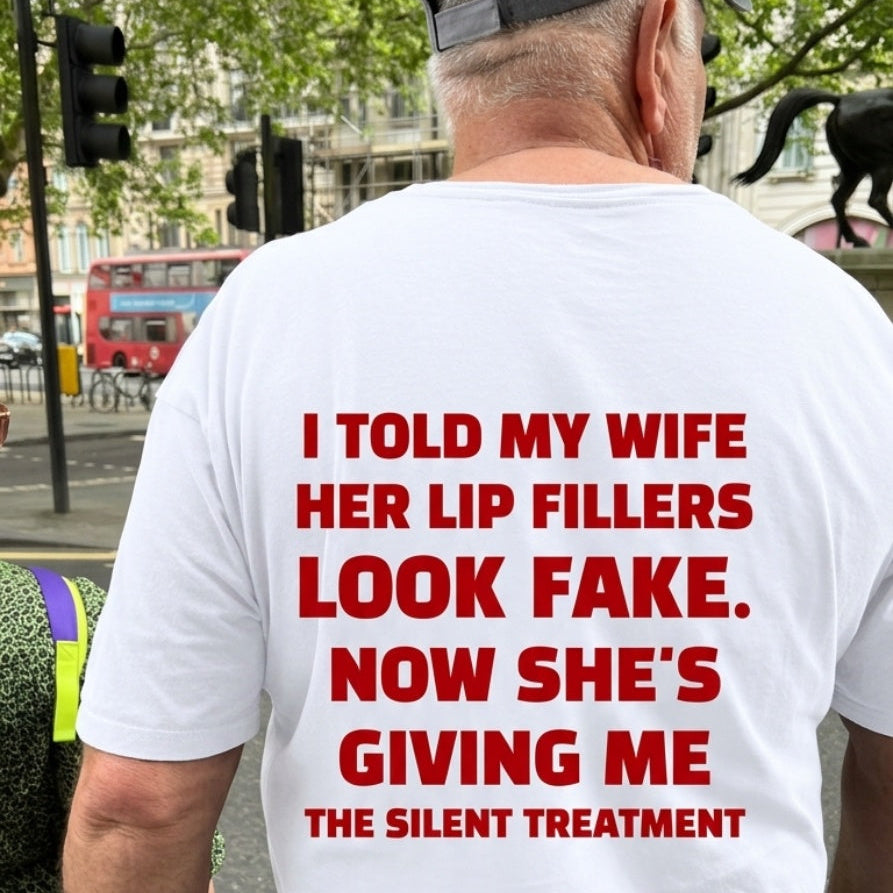 A man viewed from behind wearing a white t-shirt with bold red text printed on the back, standing on a city street. The text reads: 'I TOLD MY WIFE HER LIP FILLERS LOOK FAKE.
