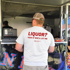 A man standing with his back to the camera in front of a food truck, wearing a white t-shirt with red text that reads 'LIQUOR? YEAH IF SHE'LL LET ME'.