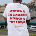 A man viewed from behind wearing a white t-shirt with bold red text that reads 'ON MY WAY TO THE GENDERLESS BATHROOM TO TAKE A SHE/IT.' and a small red logo near the bottom.