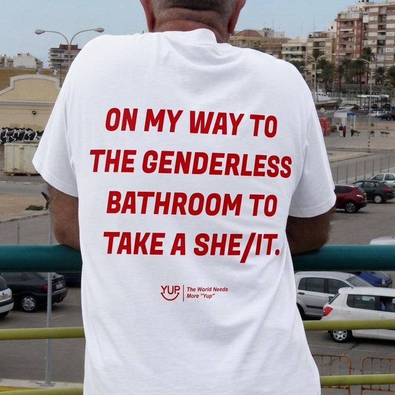 A man viewed from behind wearing a white t-shirt with bold red text that reads 'ON MY WAY TO THE GENDERLESS BATHROOM TO TAKE A SHE/IT.' and a small red logo near the bottom.