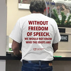 A man viewed from behind wearing a white t-shirt with bold red text that reads 'WITHOUT FREEDOM OF SPEECH. WE WOULD NOT KNOW WHO THE IDIOTS ARE' and blue jeans, standing at a counter.