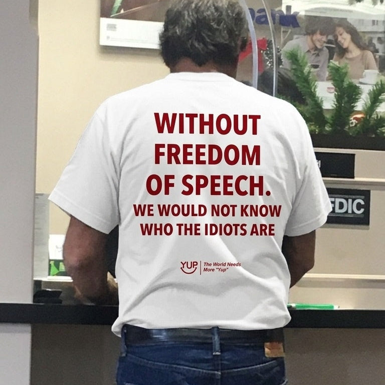 A man viewed from behind wearing a white t-shirt with bold red text that reads 'WITHOUT FREEDOM OF SPEECH. WE WOULD NOT KNOW WHO THE IDIOTS ARE' and blue jeans, standing at a counter.