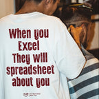 A person seen from behind wearing a white graphic T-shirt with the phrase 'When you Excel They will spreadsheet about you' printed in dark red text. The shirt is shown in a real indoor setting with another person partially visible at the side.
