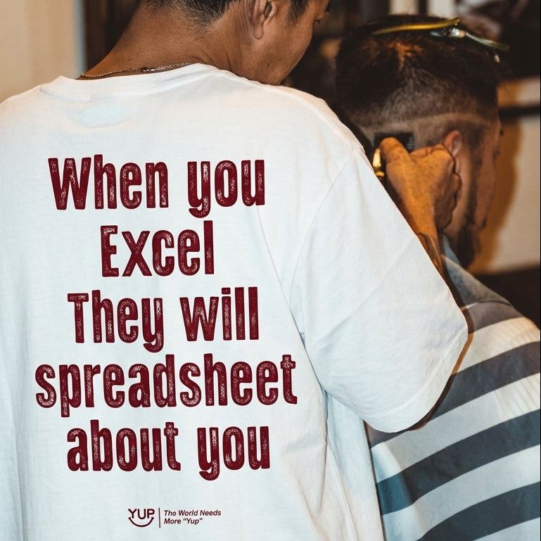 A person seen from behind wearing a white graphic T-shirt with the phrase 'When you Excel They will spreadsheet about you' printed in dark red text. The shirt is shown in a real indoor setting with another person partially visible at the side.