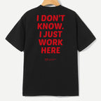 A black short-sleeve T-shirt hanging on a wooden hanger against a light gray background, featuring large red text on the back that reads 'I DON'T KNOW. I JUST WORK HERE' with a small red logo underneath.