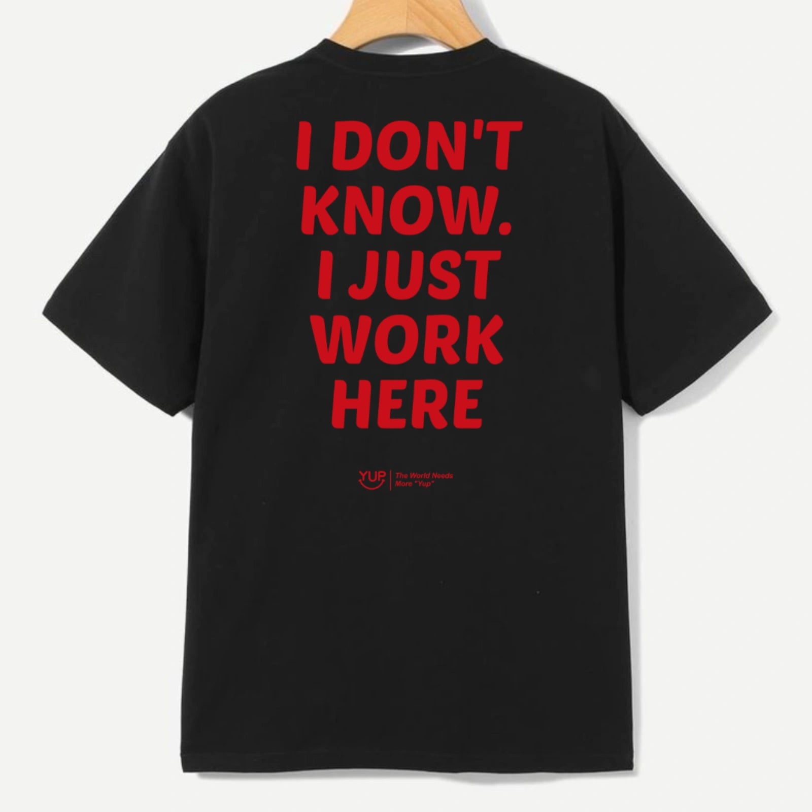 A black short-sleeve T-shirt hanging on a wooden hanger against a light gray background, featuring large red text on the back that reads 'I DON'T KNOW. I JUST WORK HERE' with a small red logo underneath.