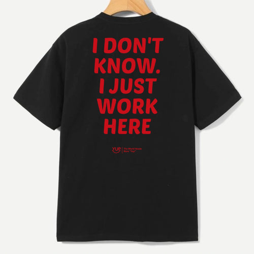 A black short-sleeve T-shirt hanging on a wooden hanger against a light gray background, featuring large red text on the back that reads 'I DON'T KNOW. I JUST WORK HERE' with a small red logo underneath.
