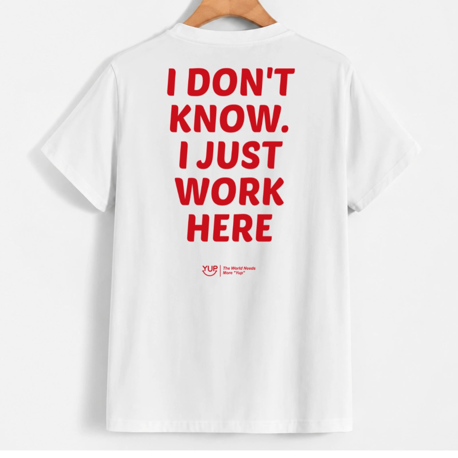 White short-sleeve T-shirt hanging on a wooden hanger, shown from the back with large red text reading 'I DON'T KNOW. I JUST WORK HERE' and a small red 'Yup' logo near the lower back.