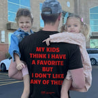 A man wearing a black graphic t-shirt with bold red text on the back, carrying two young girls in a parking lot. The shirt reads 'MY KIDS THINK I HAVE A FAVORITE BUT I DON'T LIKE ANY OF THE!'.