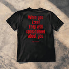 A black short-sleeve t-shirt laid flat on a textured concrete surface with dappled shadows. The back of the shirt features the phrase 'When you Excel They will spreadsheet about you' printed in a bold red font.