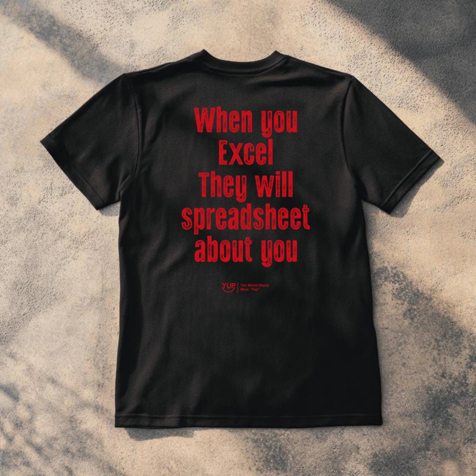 A black short-sleeve t-shirt laid flat on a textured concrete surface with dappled shadows. The back of the shirt features the phrase 'When you Excel They will spreadsheet about you' printed in a bold red font.