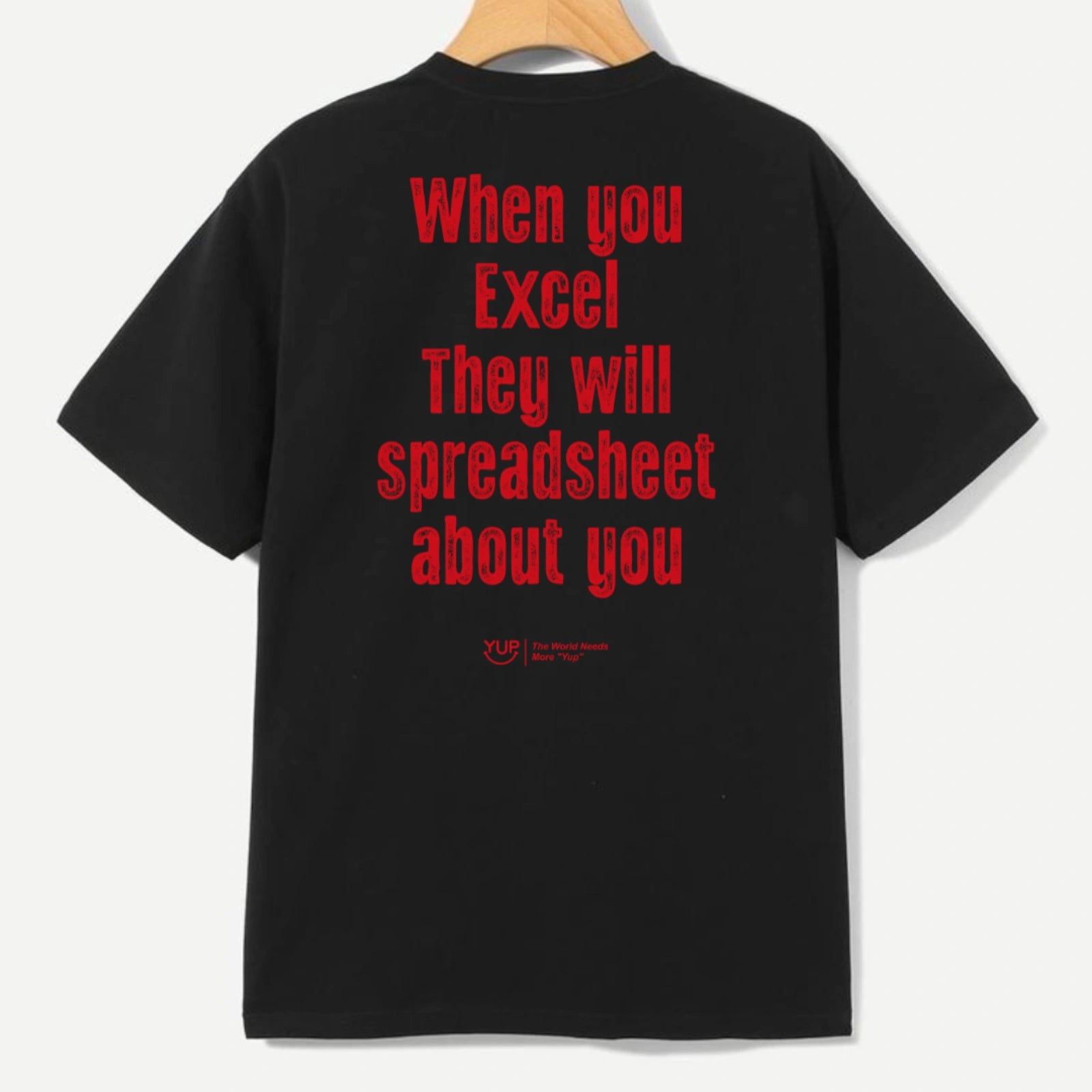 A black short-sleeve t-shirt hanging on a wooden hanger, showing the back with bold red text that reads 'When you Excel They will spreadsheet about you'.